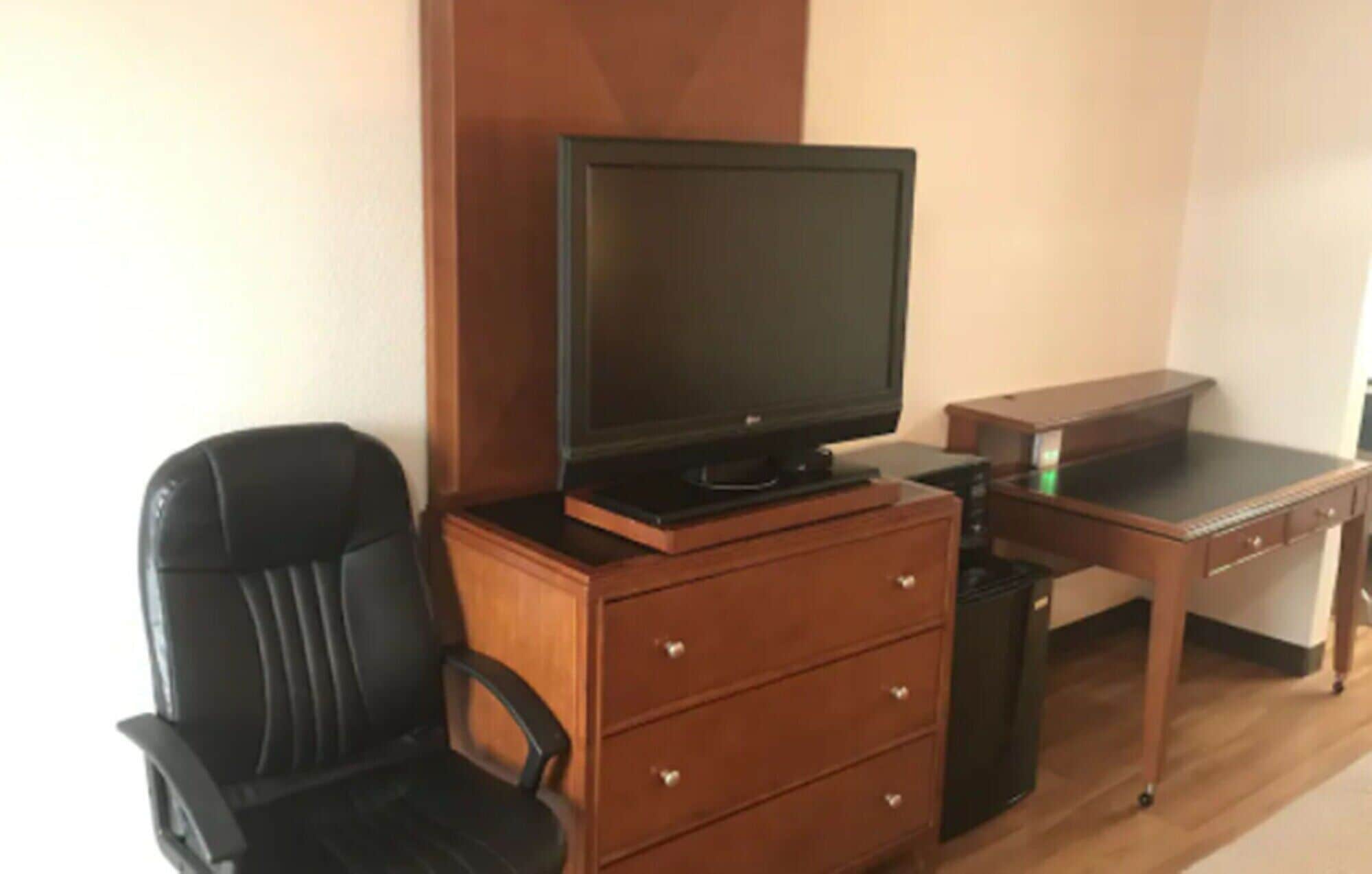 Une pièce de taille modeste, avec un bureau en bois, une chaise noire, un téléviseur posé sur un meuble en bois et une petite poubelle.