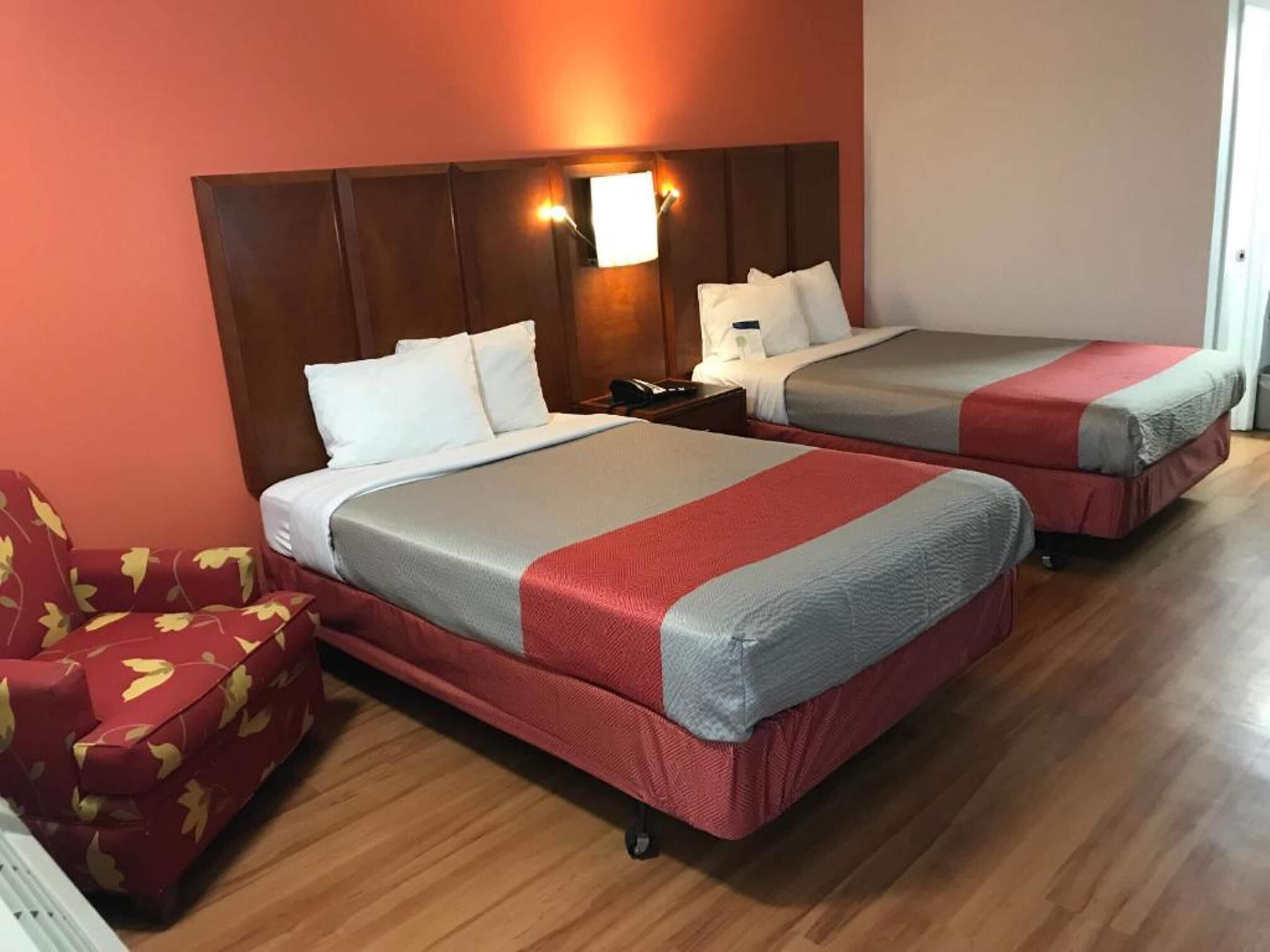 A hotel room with two beds, a wooden headboard, a nightstand, a red and yellow patterned armchair, and a red carpet.