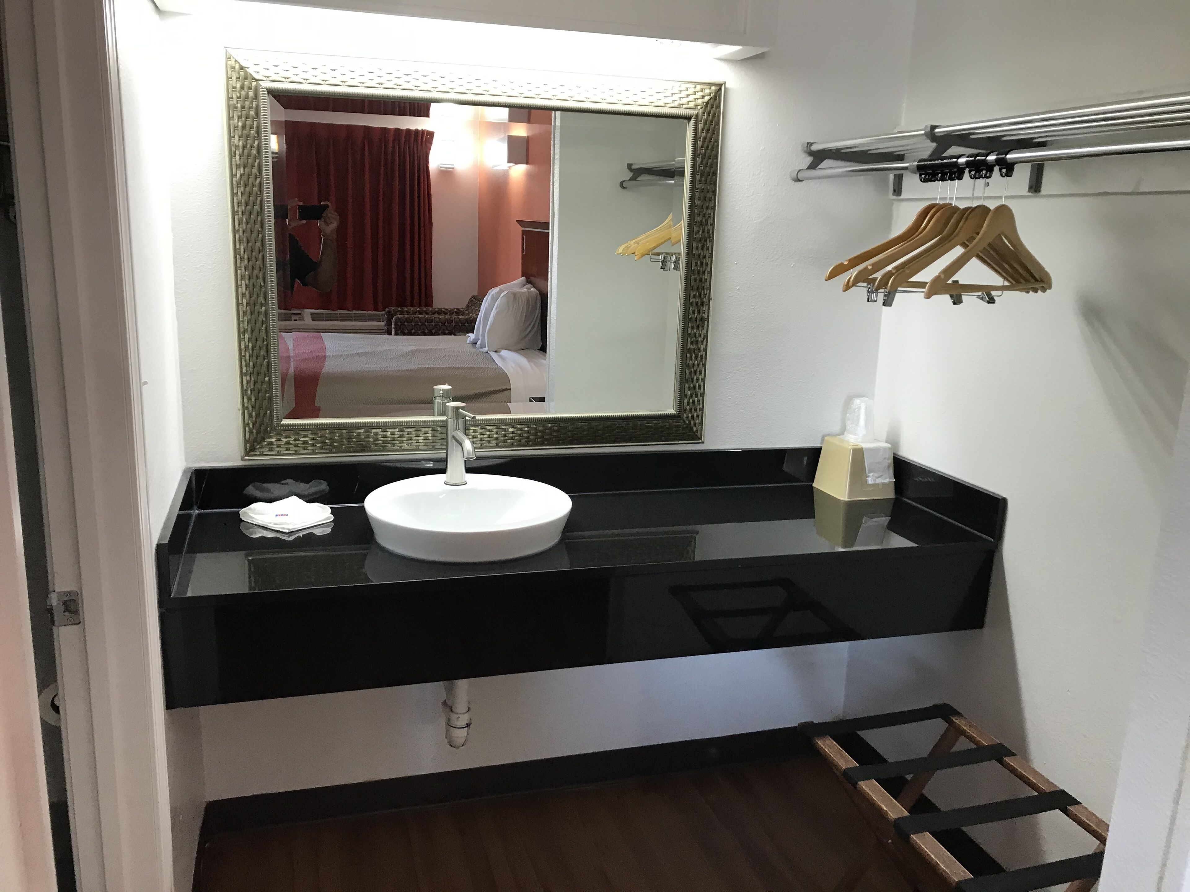 A bathroom with a round white sink, a mirror with a decorative frame, a towel rack, and a coat rack with hangers.