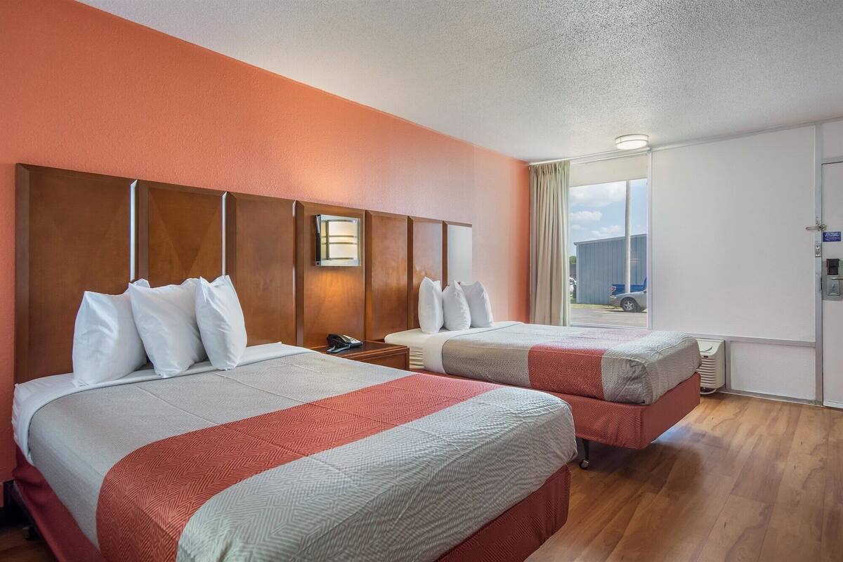 Une chambre d’hôtel avec deux lits, une tête de lit en bois, une table de chevet avec un téléphone et une fenêtre donnant sur un immeuble et une voiture.