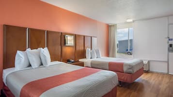Habitación de hotel con dos camas, cabecera de madera, mesita de noche con teléfono y ventana con vistas a un edificio y un coche.