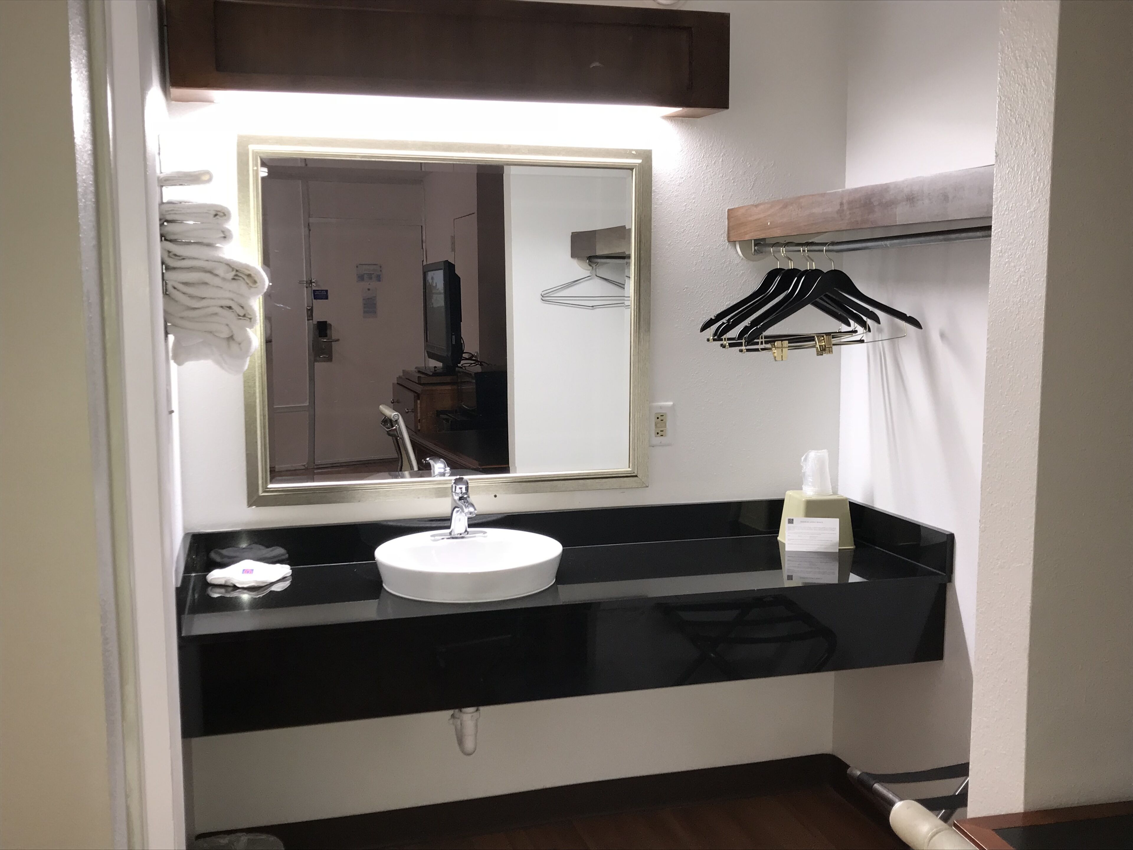 Un bagno con un lavandino bianco, un piano di lavoro nero e uno specchio sopra. Asciugamani impilati a parete e un appendiabiti con grucce nere.