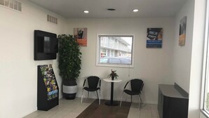 A reception area with a TV, a plant, two chairs, a small table, and a window with a view of a building. - Motel 6 Van Buren, AR (Van Buren)