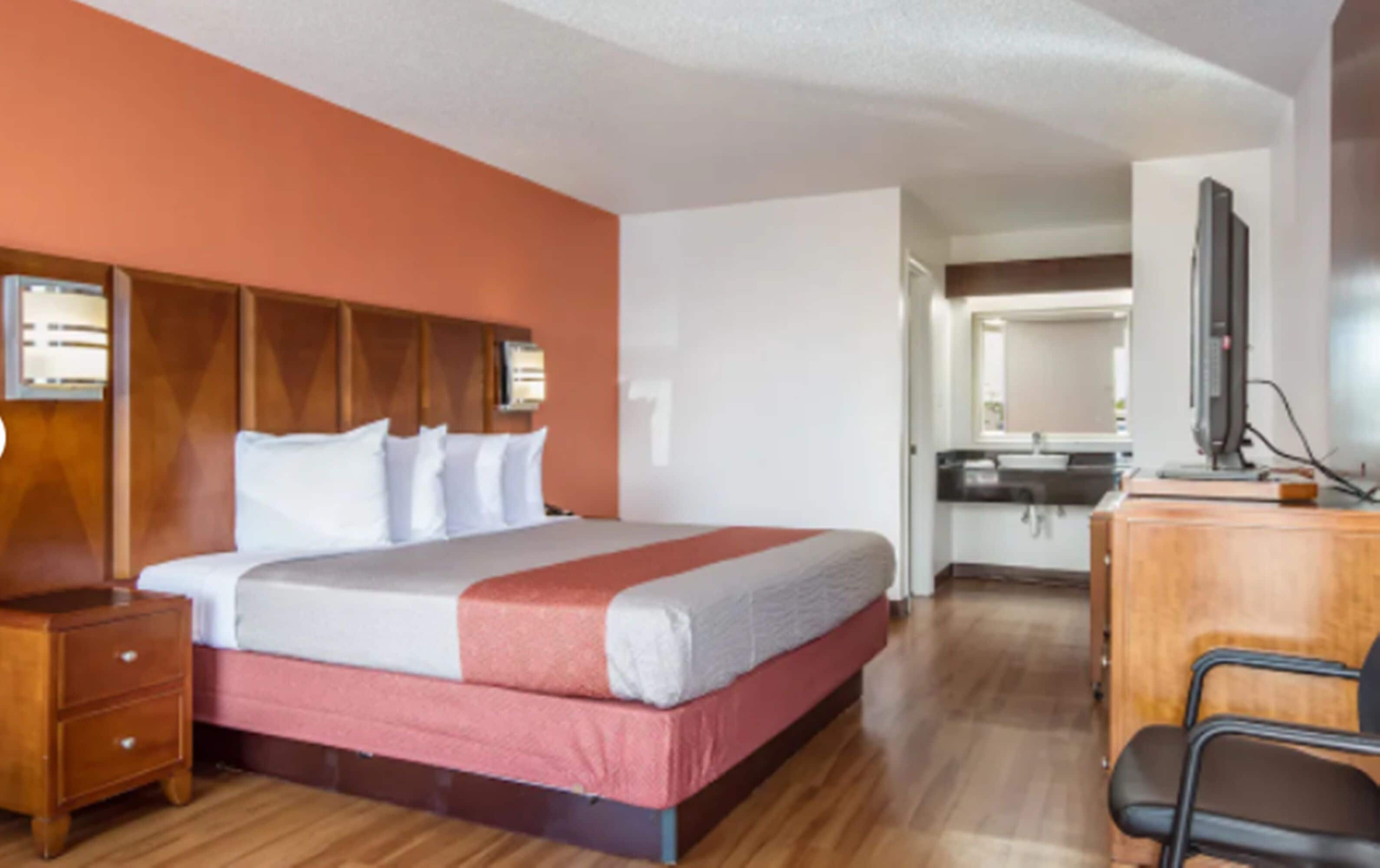A hotel room with a large bed, wooden headboard, a desk with a chair, a mirror, and a kitchenette in the background.