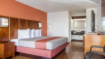 A hotel room with a large bed, wooden headboard, a desk with a chair, a mirror, and a kitchenette in the background.