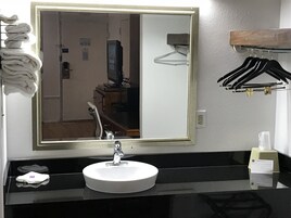 Un baño con un espejo grande, un lavabo redondo, un perchero con toallas dobladas, una encimera negra, un estante de madera con perchas y una botella pequeña sobre la encimera.