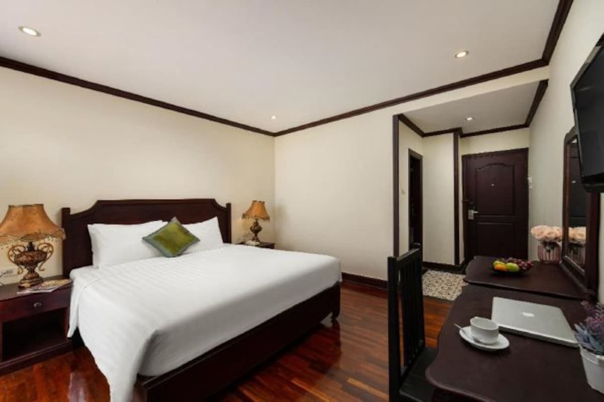 A hotel room with a wooden bed, two bedside lamps, a desk with a laptop, a cup, and a bowl of fruit.