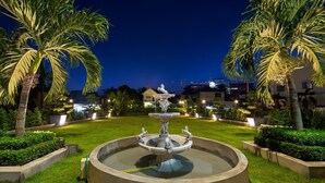A well-lit fountain with statues in a circular pool, surrounded by palm trees and well-maintained grass, with buildings in the background. - Vientiane Golden Sun Hotel & Spa (Vientiane)