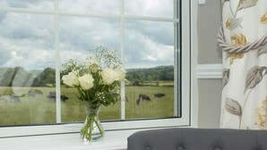 A window with a view of a field and cows, a vase with white flowers, and a grey upholstered chair. - Poole Farm (Launceston)