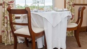 A dining table set with white tablecloth, surrounded by wooden chairs with cushioned seats, in a room with floral curtains and a view through a large window. - Poole Farm (Launceston)