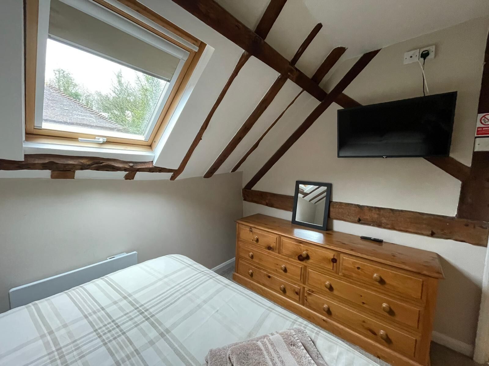 Um quarto com teto inclinado, cômoda de madeira, cama com colcha xadrez, janela com vista para árvores e TV de tela plana fixada na parede.