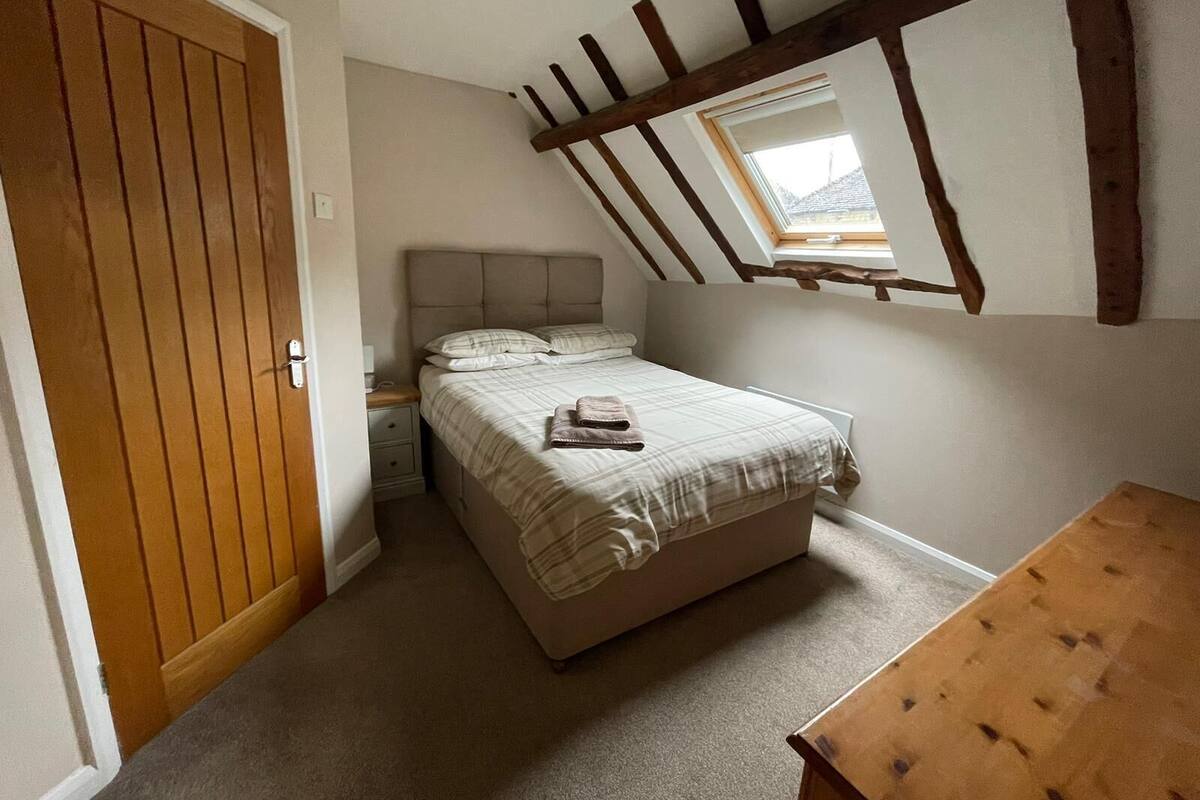 A bedroom with a sloped ceiling, a bed with a headboard, a nightstand, a wooden door, and a small window.