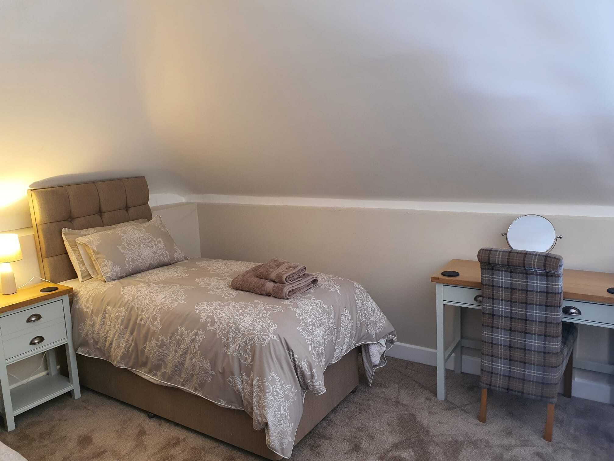 A neatly made bed with a headboard, a bedside table with a lamp, a chair with a mirror, and a small drawer unit.
