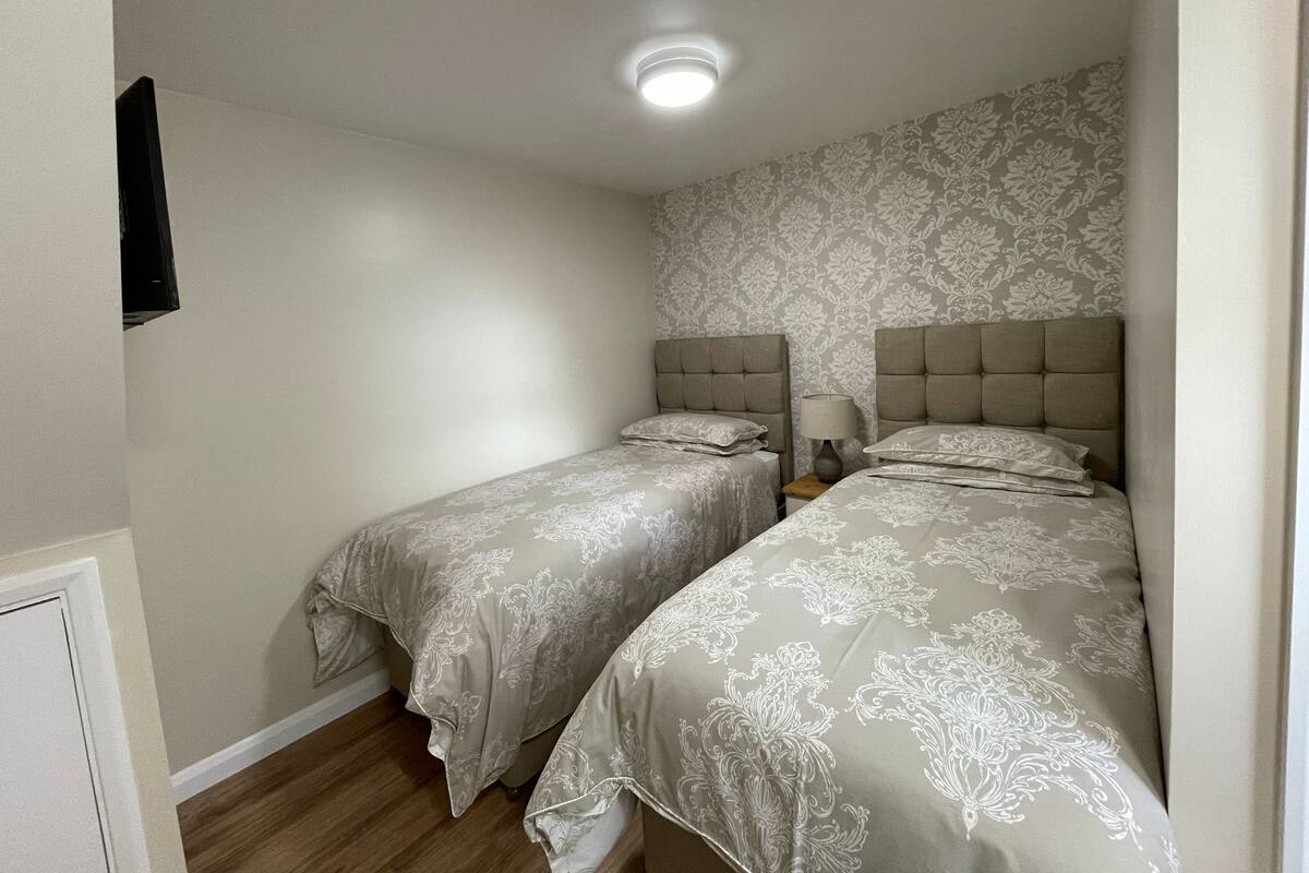 A small, compact bedroom with two single beds, a nightstand with a lamp, and a ceiling light.