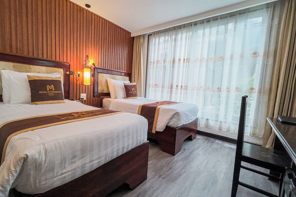 A hotel room with two beds, wooden headboards, a large window with sheer curtains, and a wooden nightstand.
