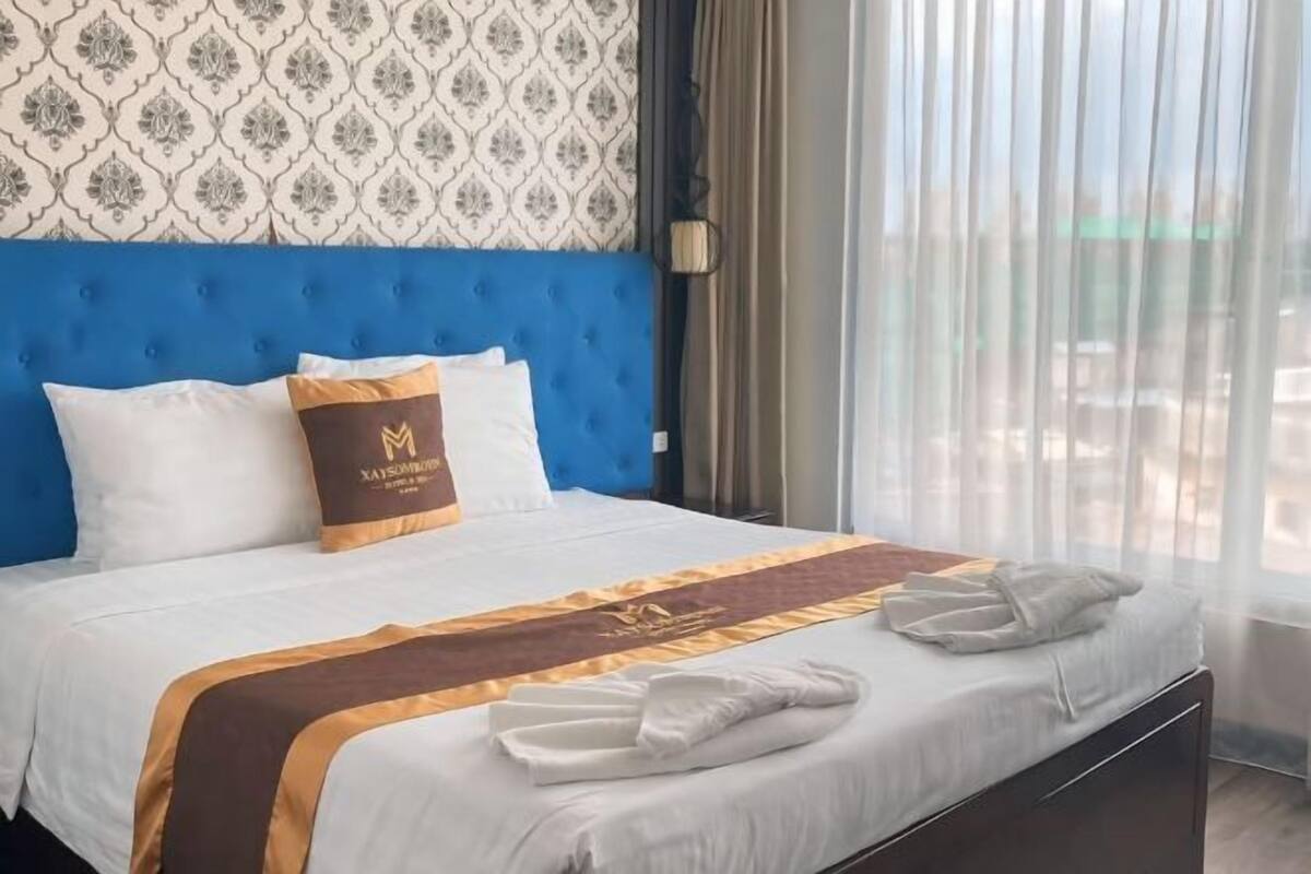 A hotel room with a blue headboard, a bed with white linens, a brown throw blanket, and a pillow with a logo.