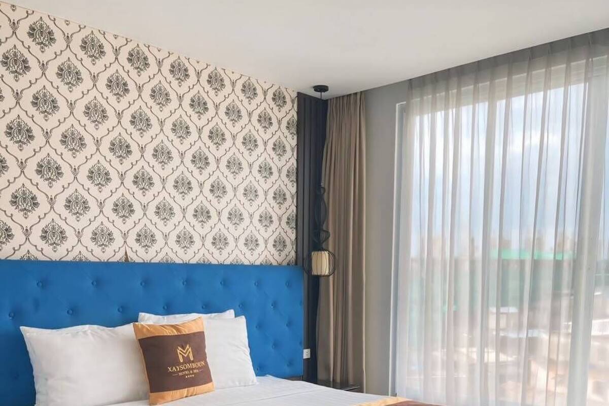 A hotel room with a large bed, a blue headboard, a patterned wall, and a view through sheer curtains.