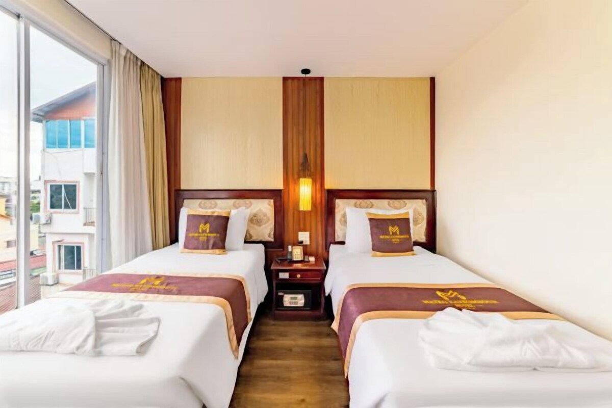 A hotel room with two beds, a nightstand, a lamp, and a window with curtains.