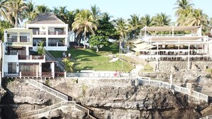 A coastal hotel with a rocky cliff, a staircase leading to the beach, and palm trees in the background.