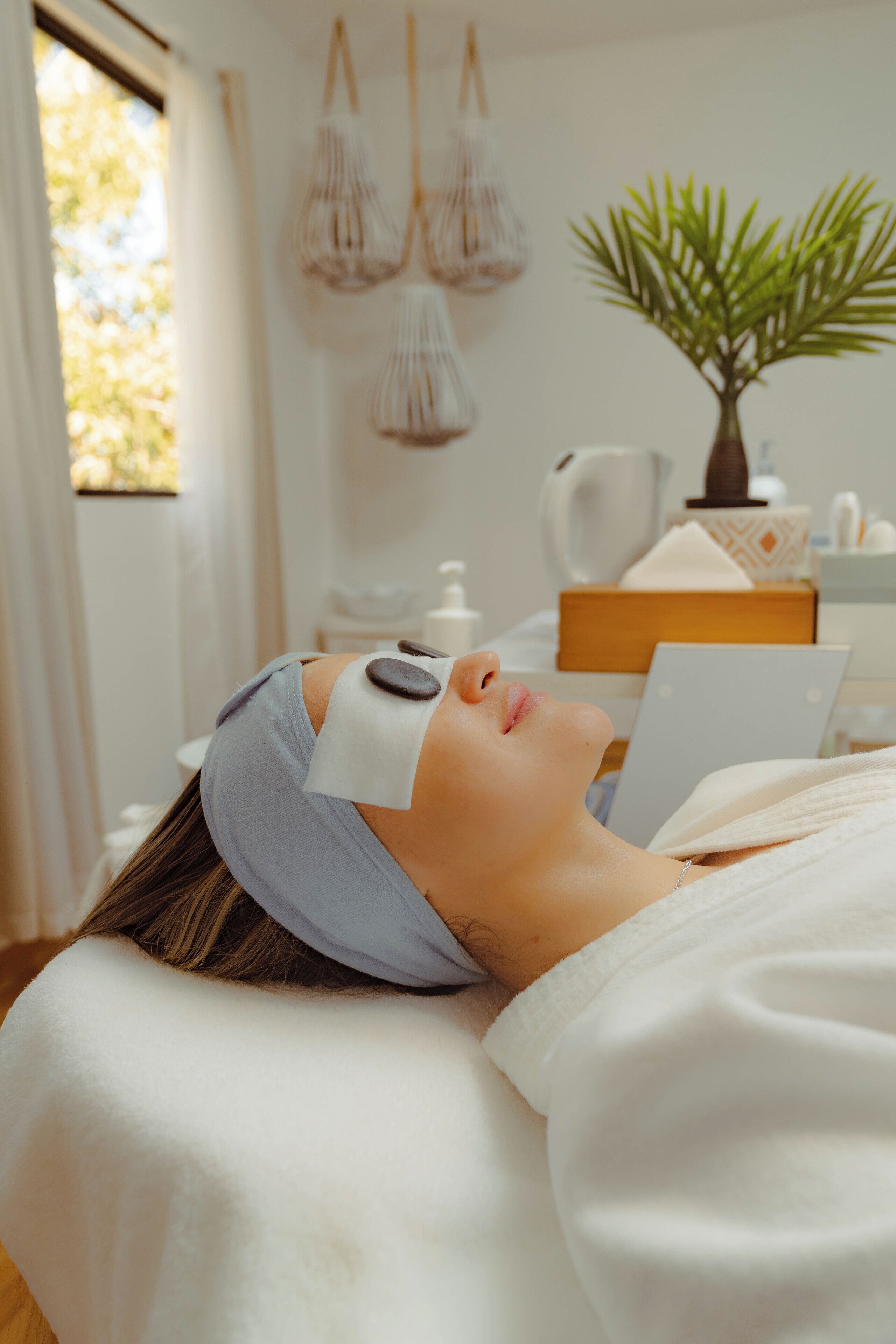 A person lying down with a headband and a white towel, in a room with a plant, a kettle, and a box on the table.