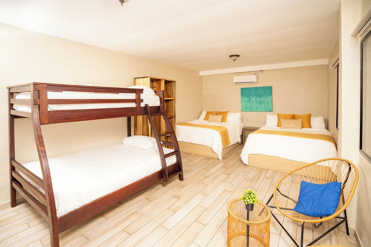 A room with bunk beds, a single bed, a small table with a plant, a wicker chair, and a painting on the wall.