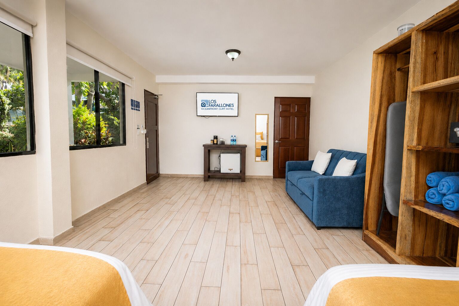 Een hotelkamer met een houten vloer, een blauwe bank, een houten kledingkast en een bureau met spiegel.