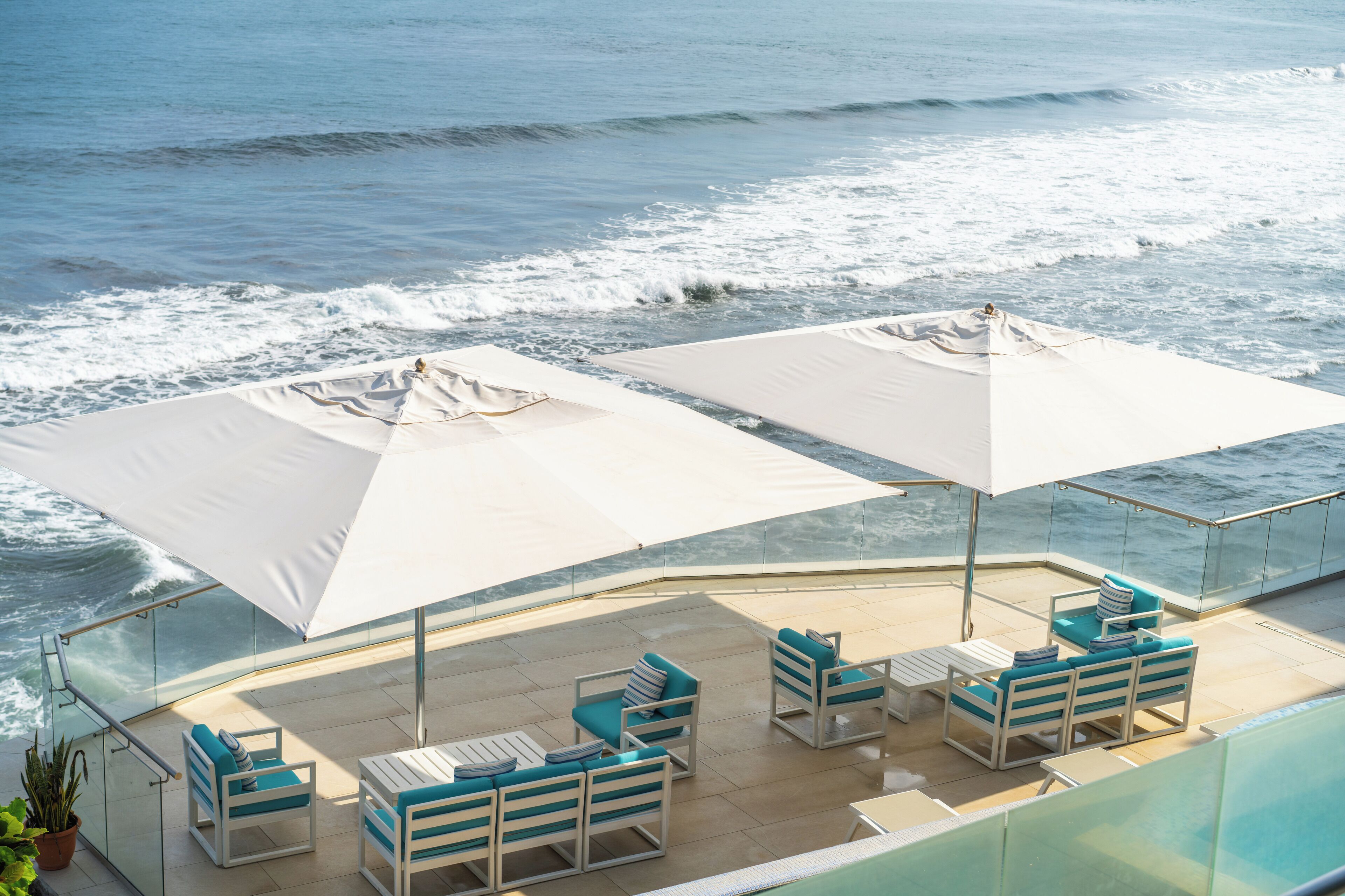 A sunlit outdoor seating area with white umbrellas, blue cushions, and a glass balustrade overlooking the ocean.