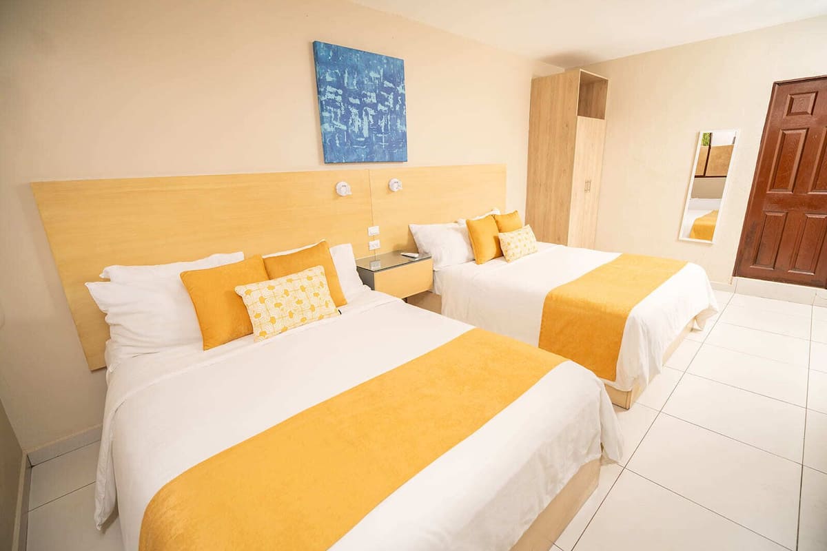 Habitación de hotel con dos camas, cada una con ropa de cama blanca y amarilla, un cabecero de madera, un cuadro abstracto azul en la pared y un armario de madera.