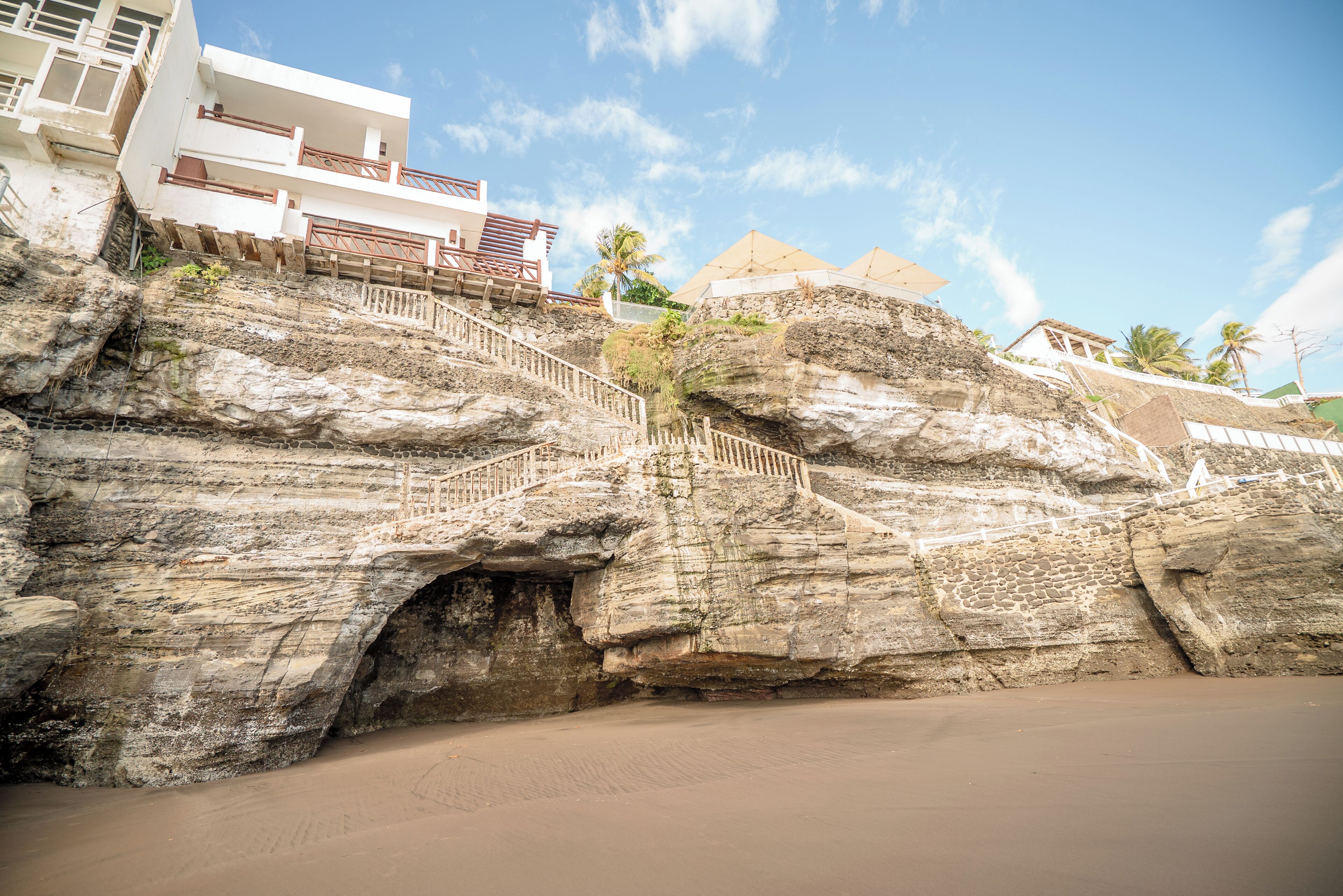 A coastal hotel with a staircase leading to a beach, a palm tree, and a clear sky.