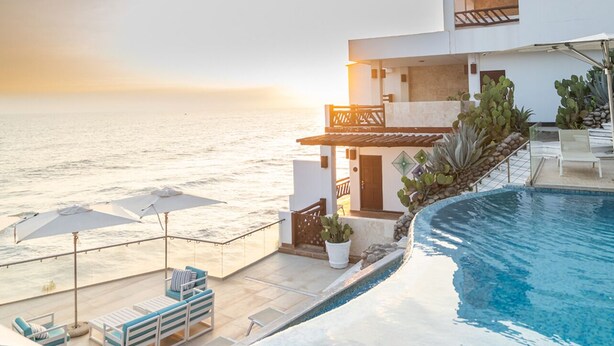 A modern hotel with a pool overlooking the sea, featuring a terrace with sun loungers and umbrellas.