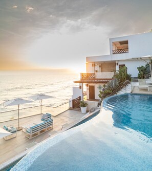 A modern hotel with a pool overlooking the sea, featuring a terrace with sun loungers and umbrellas.