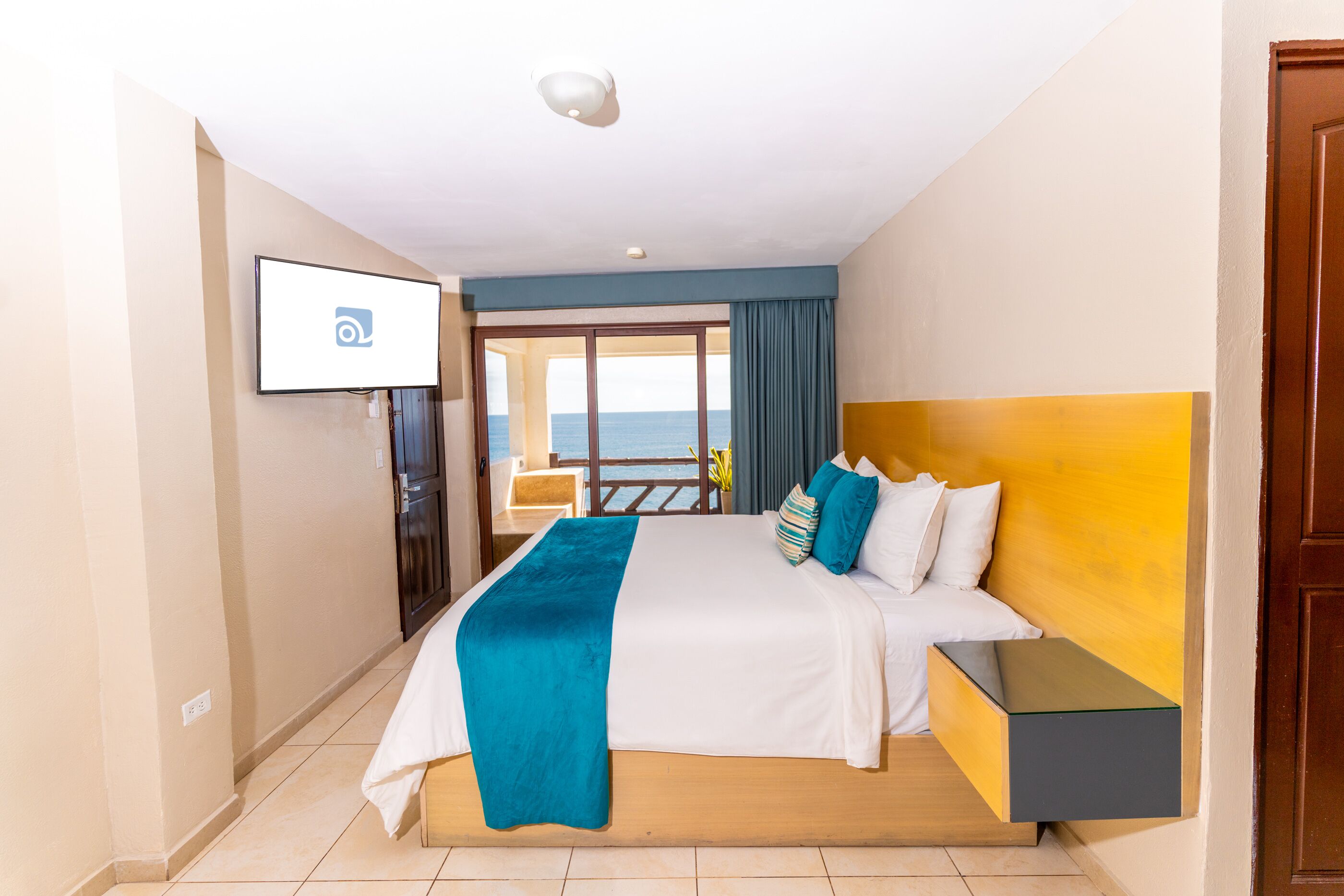 A hotel room with a bed, a TV, a nightstand, and a view of the sea through a balcony door.