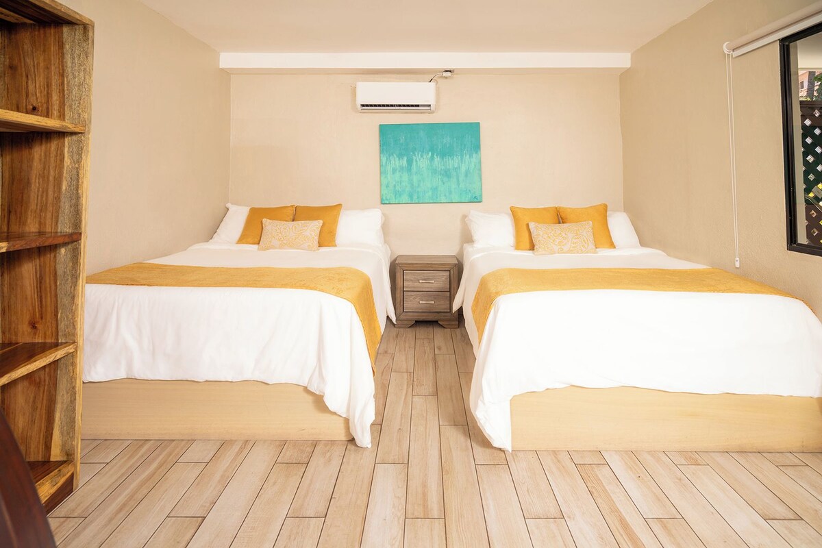 A hotel room with two beds, a wooden bookshelf, a small wooden nightstand, and a teal painting on the wall.