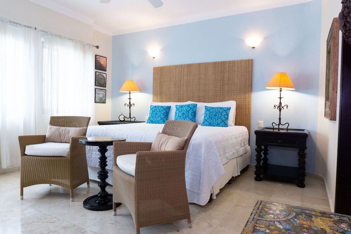 Habitación de hotel con una cama, dos sillones de mimbre, una mesita, dos lámparas y un tapete estampado.