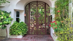 A gated entrance with a decorative metal gate, a brick pathway, and lush greenery including flowering plants. - Royal Palms by Bric (Playa del Carmen)
