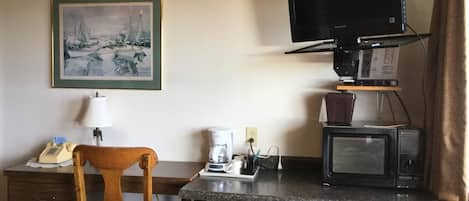 A compact hotel room with a kitchenette, a wooden chair, a framed painting, a television, and a microwave.