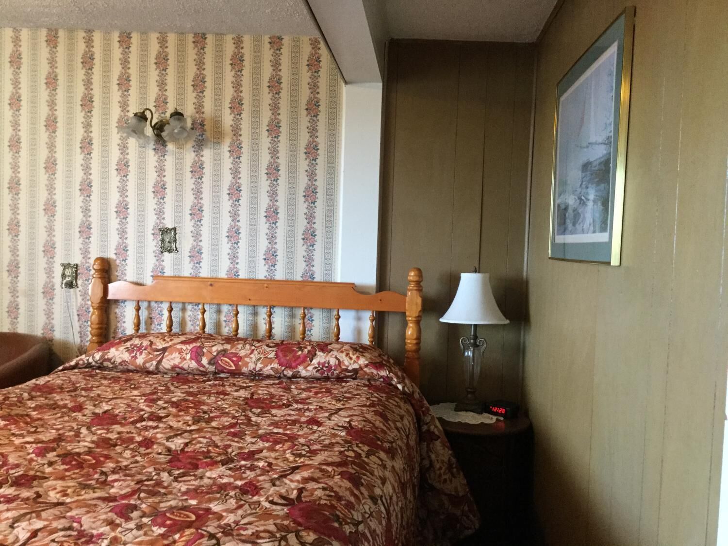 A hotel room with a wooden bed, floral-patterned bedspread, a nightstand with a lamp, and a framed picture on the wall.