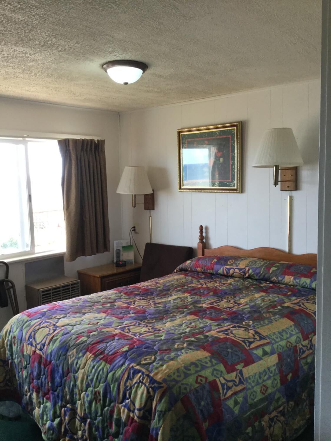 A hotel room with a large bed, a bedside table, a lamp, a picture frame, and a window with curtains.