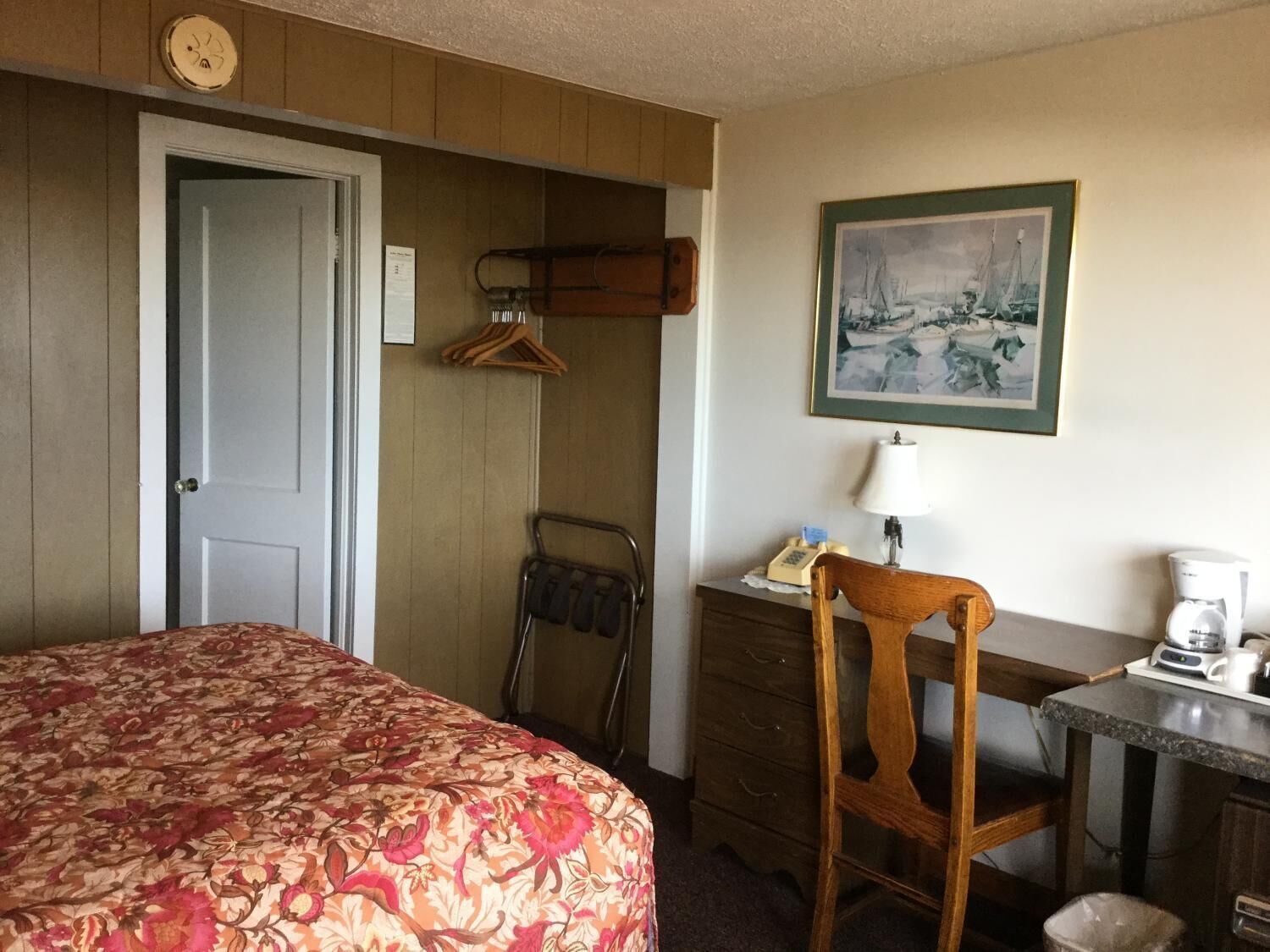 A hotel room with a bed, a wooden chair, a desk with a lamp, a coffee maker, and a framed picture on the wall.