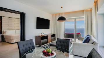A modern living room with a glass table, a sofa, a TV, and a view of the sea.