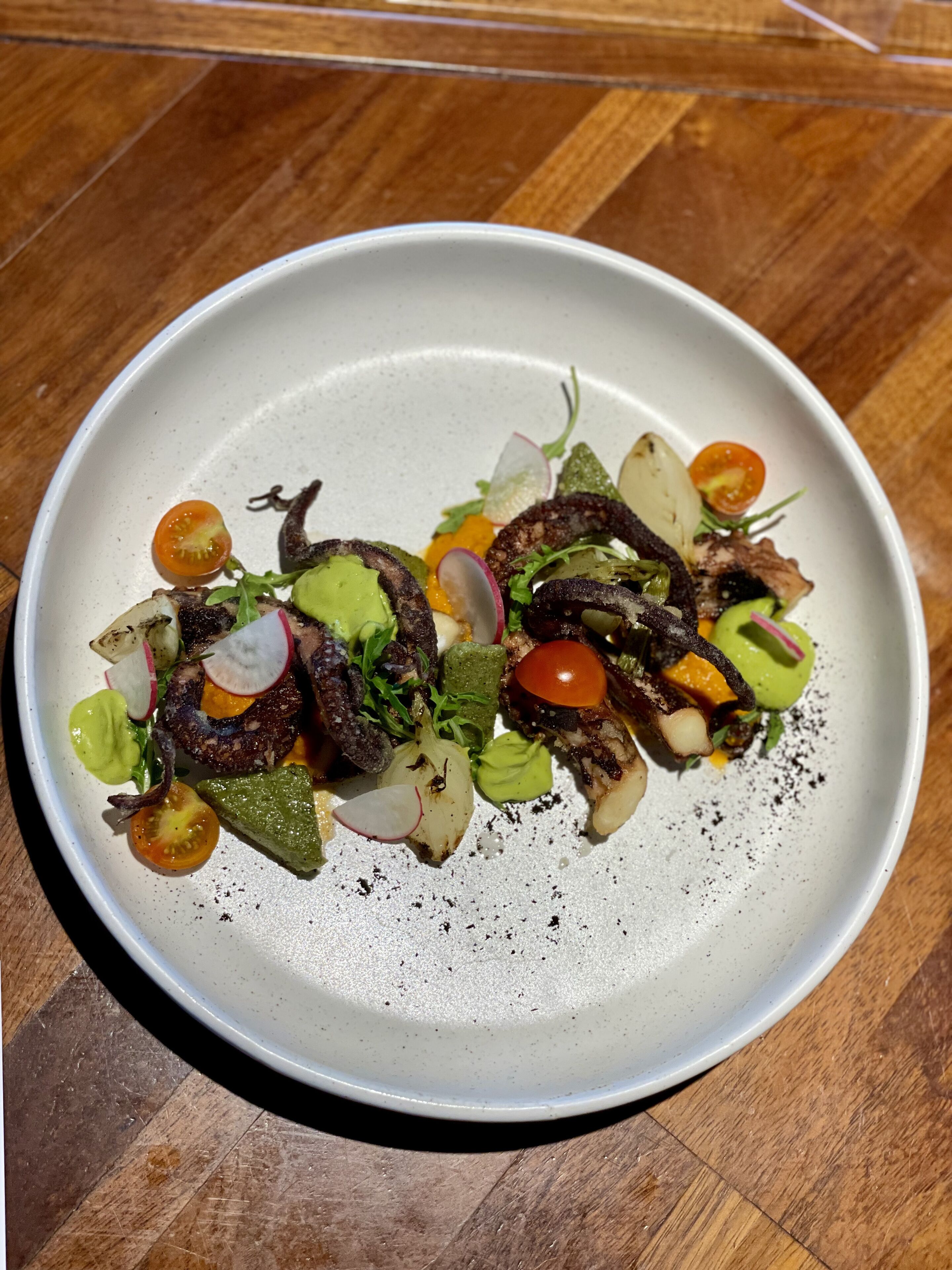 A white plate with a variety of food items including vegetables and possibly meat, arranged on a wooden table.