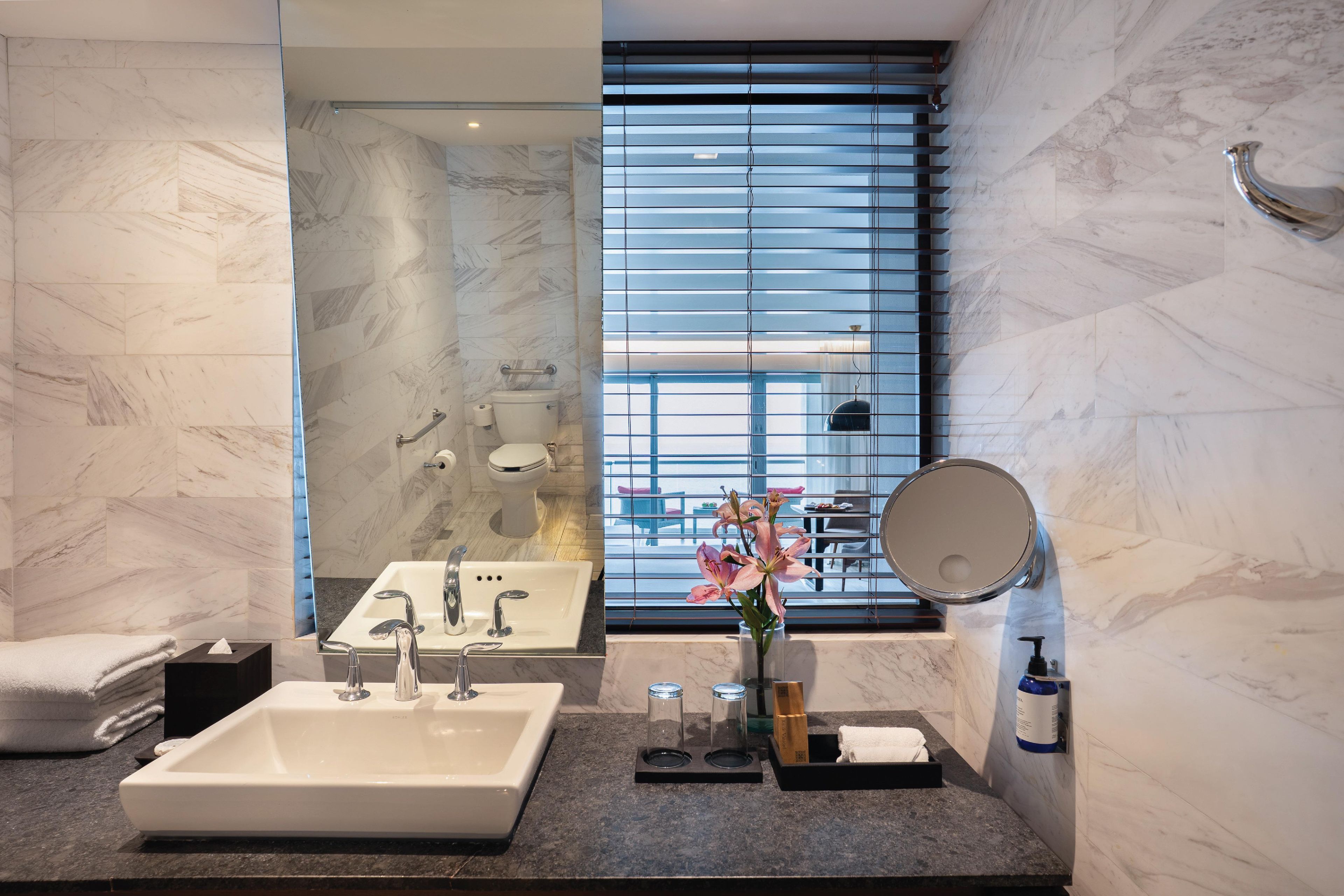 Un baño moderno con un lavamanos amplio, espejo y ventana con persianas.