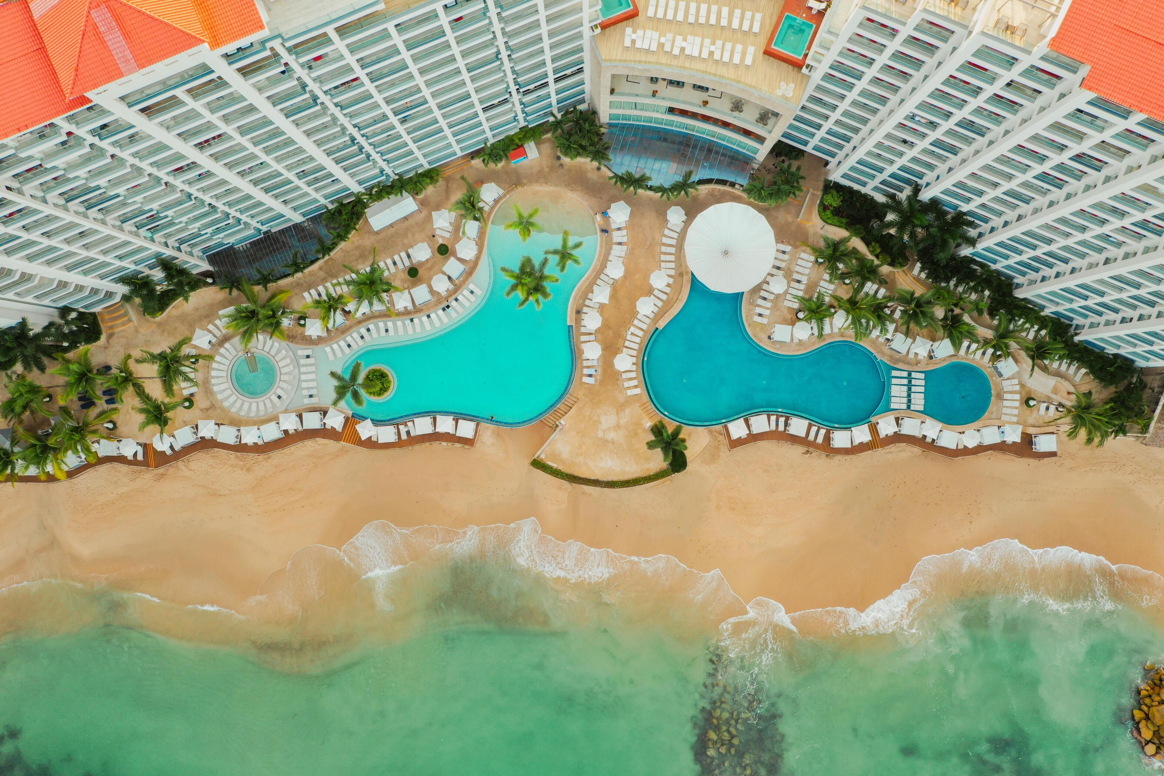 An aerial view of a resort with multiple swimming pools, a beach, and high-rise buildings.