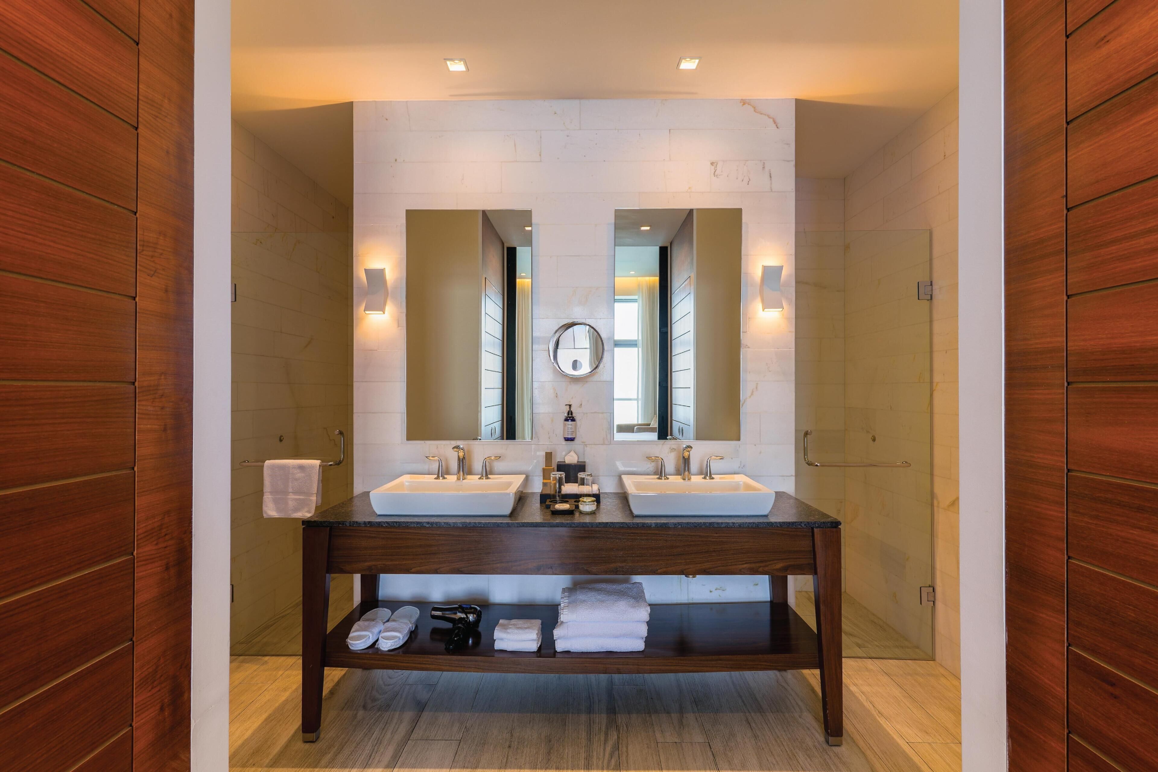 A bathroom with two sinks, a mirror, and a shelf with towels and toiletries.