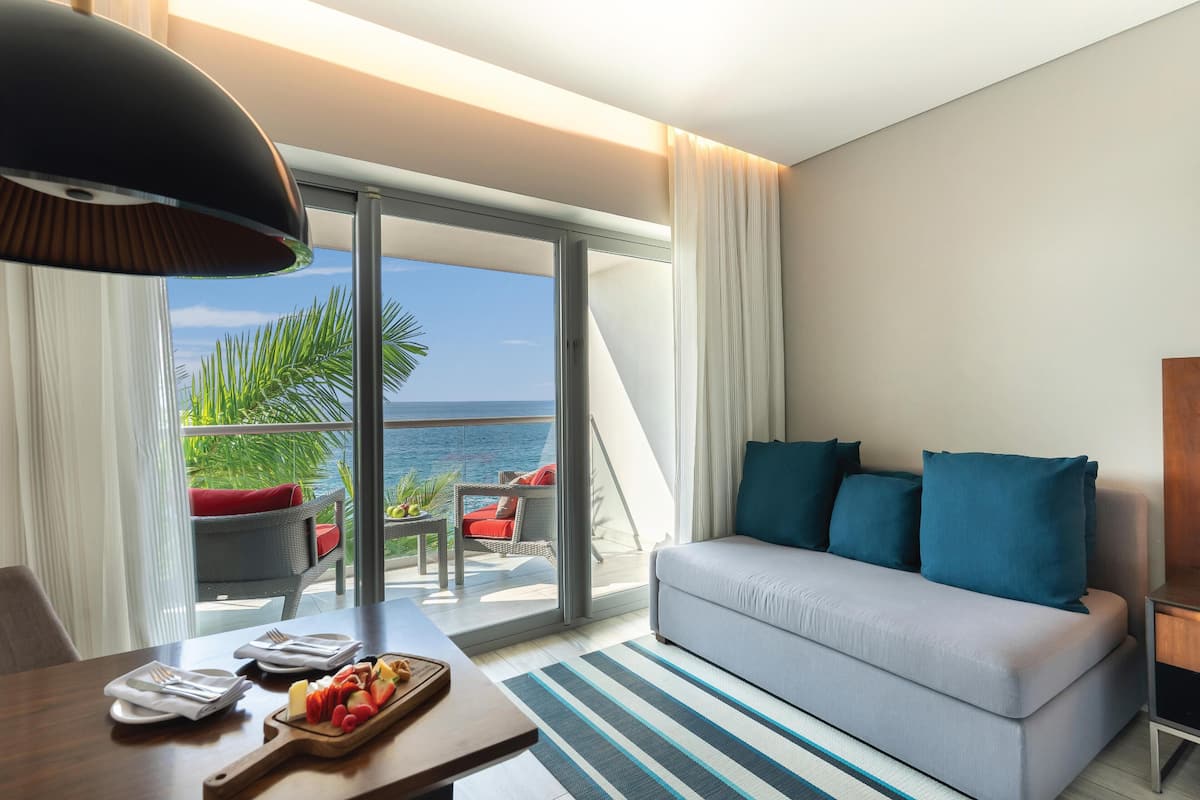 A hotel room with a balcony, a sofa, a dining table with a fruit platter, and a view of the sea.