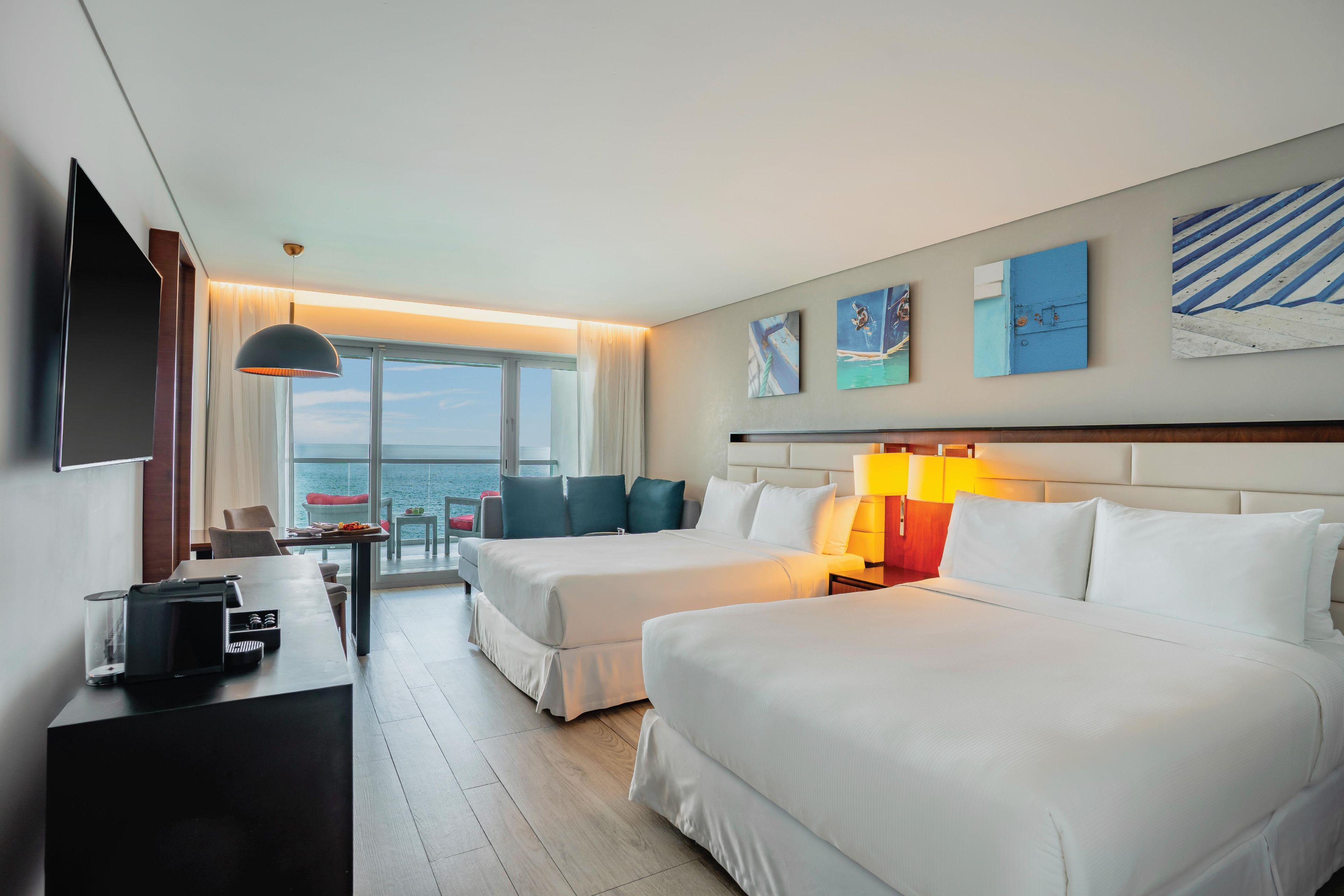 A hotel room with two beds, a desk, a TV, and artwork on the walls.