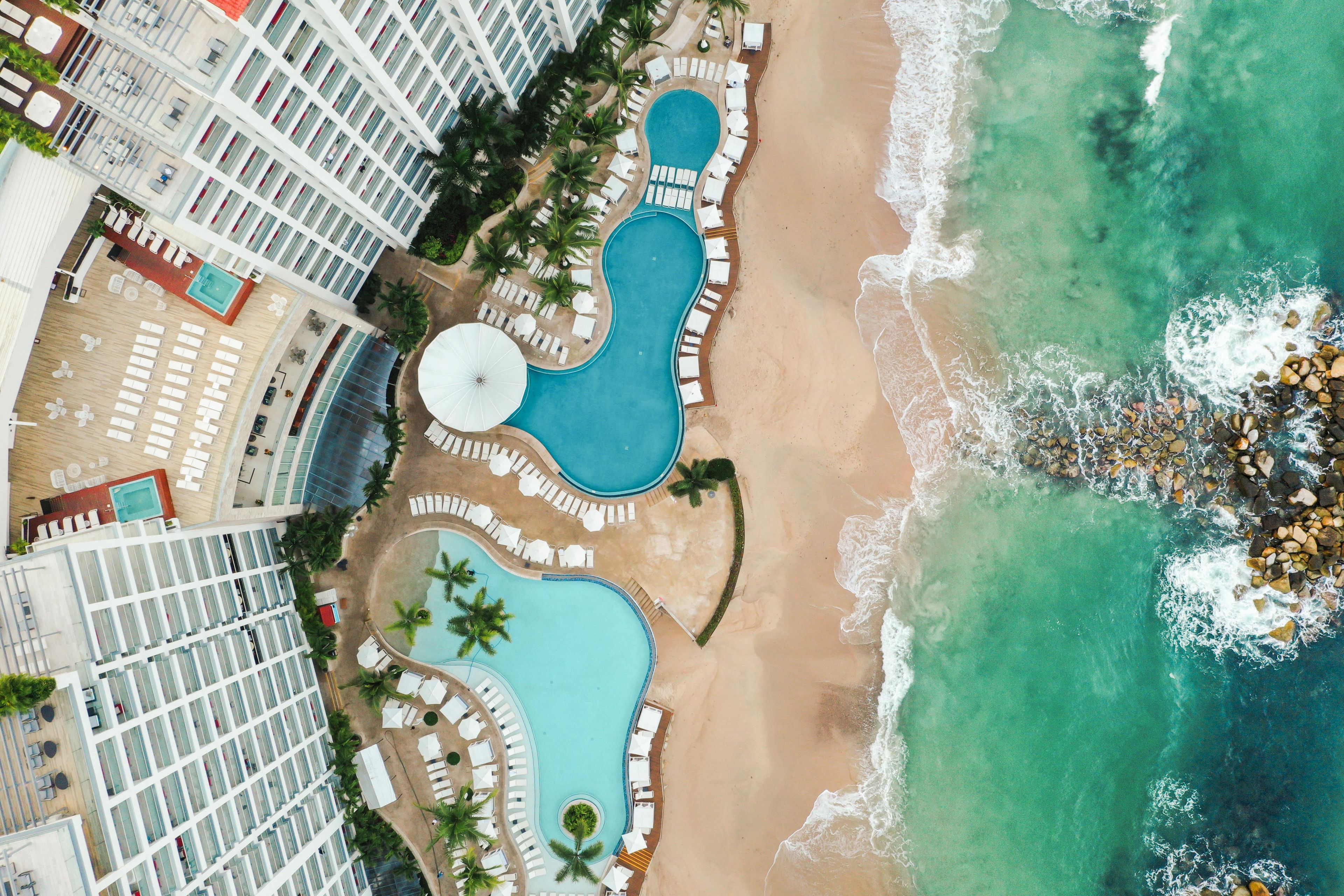Eine Vogelperspektive zeigt einen luxuriösen Urlaubsort mit mehreren Schwimmbecken, einem Strand und einem Hotel mit Balkonen.