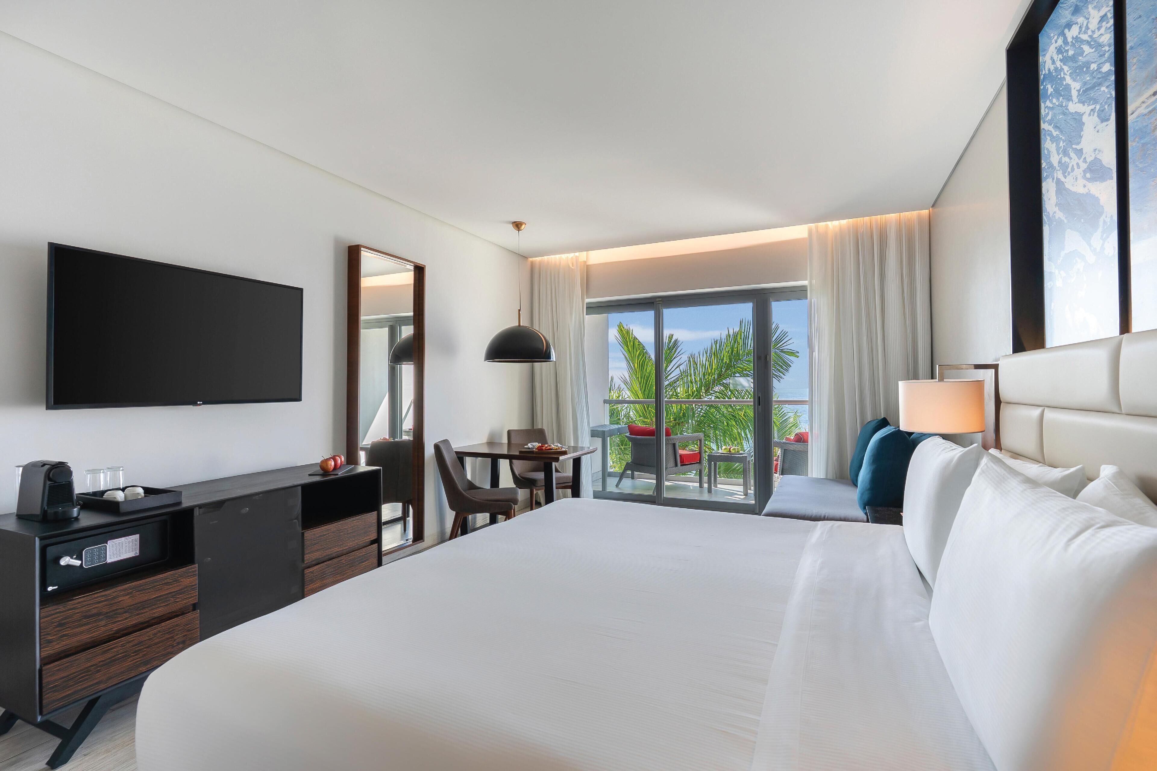 A hotel room with a large bed, a desk with a flat-screen TV, a chair, a small table, and a balcony with a view.