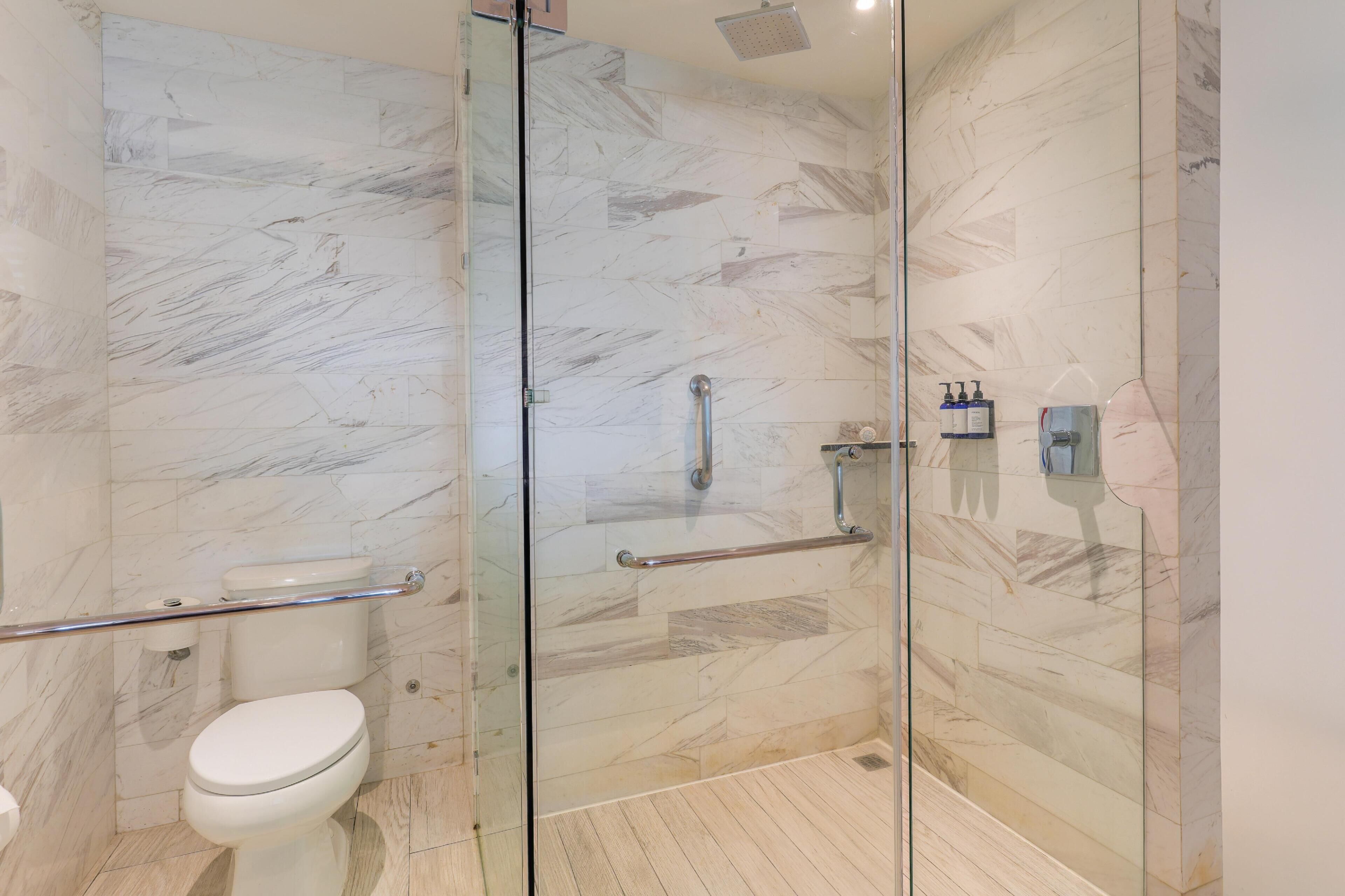 A modern bathroom with a glass shower enclosure, marble walls, and a toilet with a grab bar.