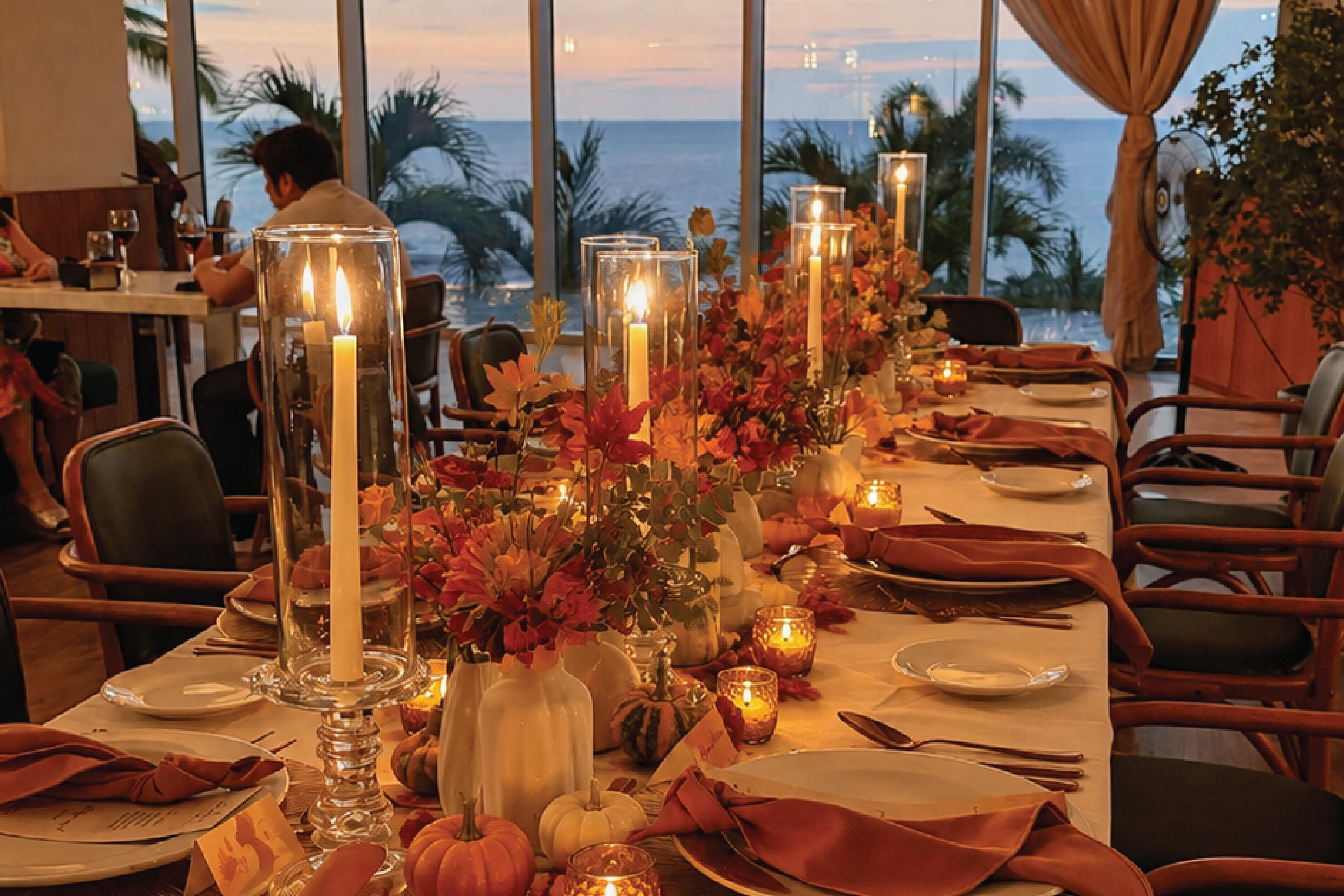 A dining table set for a meal with candles, autumn leaves, and a view of the ocean.